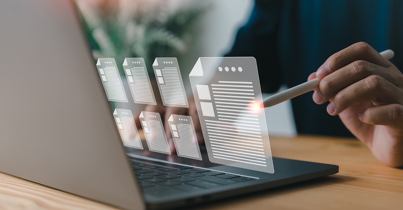 A person's hand holding a pen over a laptop screen displaying a document with text and icons.