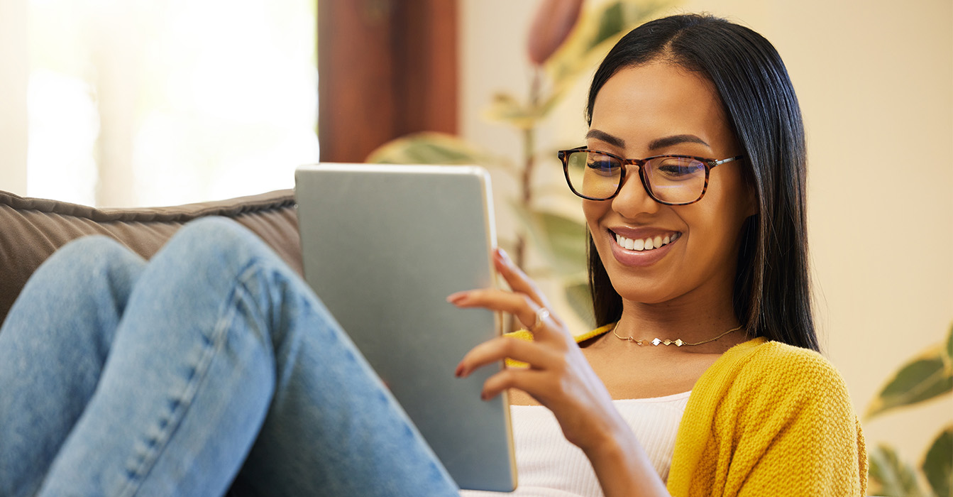 A woman with glasses is sitting on a couch, smiling, and looking at her tablet computer while holding a beverage.