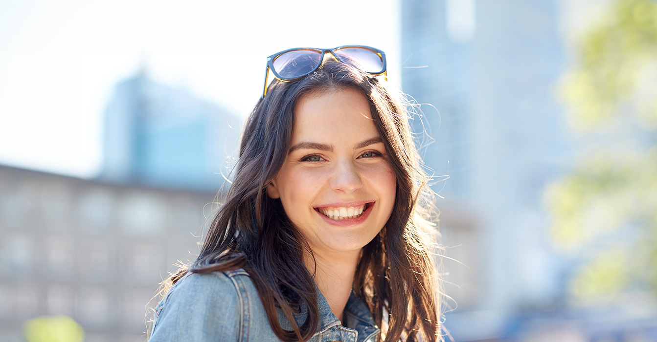 The image shows a smiling woman wearing sunglasses and a denim jacket, standing outdoors during daylight.