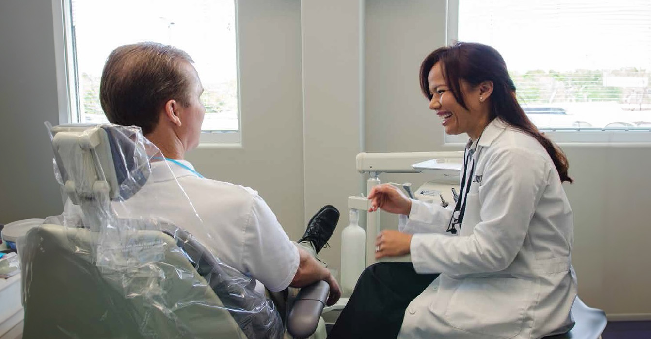 The image shows a dental office setting where a dentist is examining a patient s teeth while sitting on a chair with wheels, assisted by another person who appears to be a dental hygienist standing behind them.