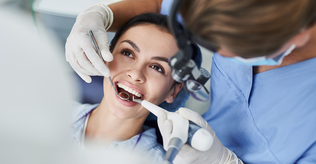 The image shows a woman sitting in a dental chair with her mouth open, receiving dental care from a professional wearing surgical gloves and a face mask.