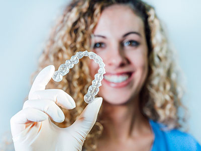 A woman holds up a clear plastic dental retainer, smiling at the camera.