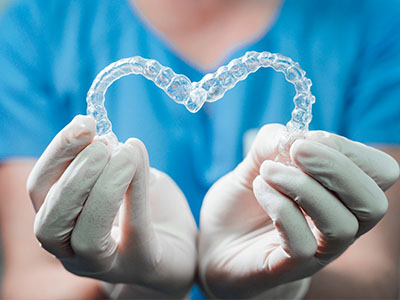 The image shows a person s hands holding up a heart-shaped object with a transparent appearance, resembling a dental impression, against a background that suggests a medical or dental setting.
