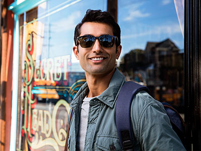 The image shows a man standing outside a shop window, wearing sunglasses, a backpack, and a jacket, with a smile on his face.