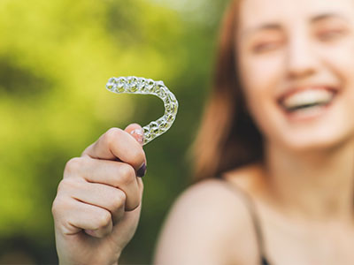 An alternative text description  A smiling woman holding a toothbrush with a smiley face on it, set against a blurred background of greenery.