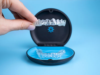 The image depicts a person s hand holding a clear plastic tray with a blue lid containing an impression of a set of teeth, placed next to a box labeled  Teeth Impressions.