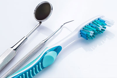 The image shows a collection of dental tools including a toothbrush with blue bristles, a pair of scissors, a small spoon-like instrument, and other utensils typically used in dentistry, arranged on a white surface.