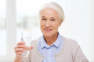 The image shows an elderly woman holding a glass of water and smiling at the camera.