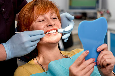 The image shows a person sitting in a dental chair with a large blue tooth model held up to their mouth by a dental professional, who is adjusting the model with gloved hands while the patient smiles at the camera.