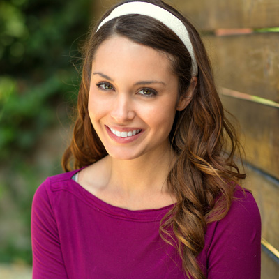 A young woman with long brown hair wearing a purple top and a white headband, smiling at the camera.