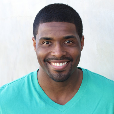 This is a color photograph featuring an older couple sitting closely together with smiles on their faces, posing for the camera. The man has gray hair and mustache, while the woman has short, light-colored hair and wears glasses. They are outdoors, possibly in a park or garden setting, as suggested by the natural lighting and greenery visible in the background. Both individuals wear casual clothing, with the man wearing a patterned shirt and the woman in a scarf. The overall mood of the image is warm and affectionate.