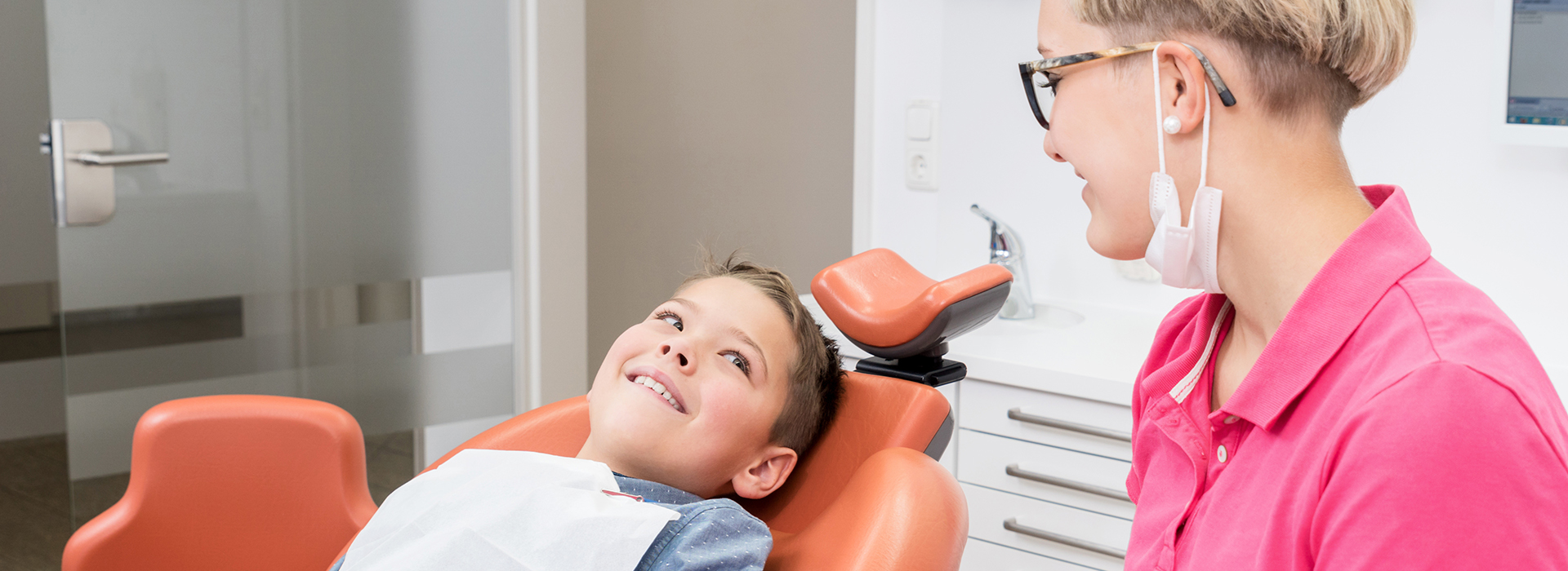 A dentist s office with a young child sitting in the dental chair, smiling at the camera, while a woman, presumably the dentist, smiles at the child from behind.