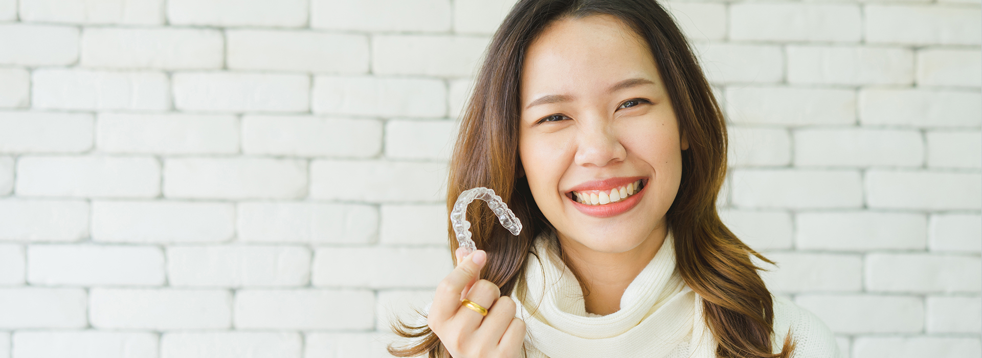 A young woman with a bright smile holds up a toothbrush, standing against a brick wall background.