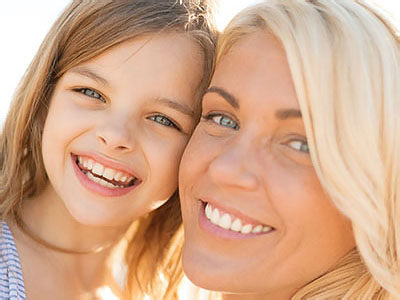 This is a portrait-style photograph featuring a smiling woman with light skin and blonde hair, wearing a blue top, and a young child with dark hair, both looking at the camera. They appear to be outdoors during daylight, and the woman s gaze is directed towards the child. The background is blurred but suggests an outdoor setting with natural lighting.