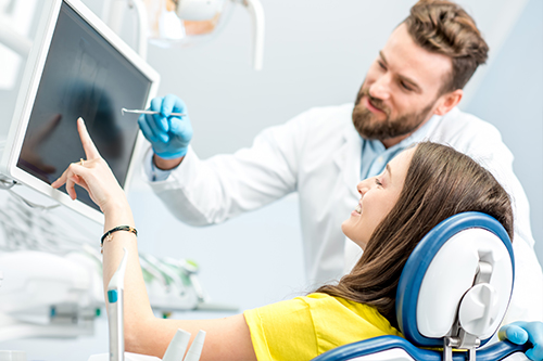 In the image, there is a man in a white lab coat standing next to a woman who is sitting in a dental chair with her head resting on a chin rest, both of them are in front of a large computer screen and monitor setup.