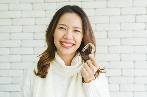 The image shows a woman holding a white object with her hand, smiling at the camera, against a brick wall background.